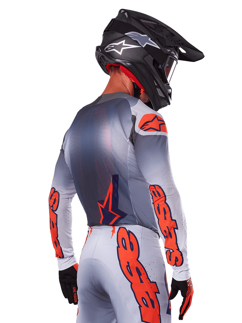 Piloto de motocross desde atrás con el Jersey Alpinestars 2025 Supertech Lipan en Gris Naranja Fluo, con logotipos naranjas llamativos en las mangas y la espalda, combinado con pantalones a juego y casco negro, equipamiento de carreras profesional para rendimiento off-road
