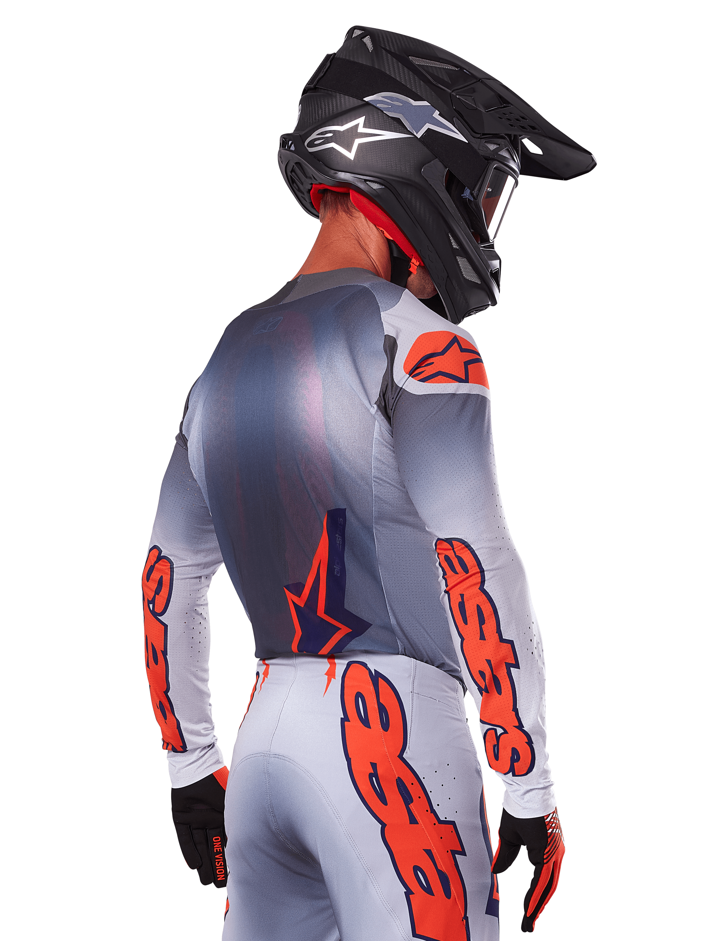 Piloto de motocross desde atrás con el Jersey Alpinestars 2025 Supertech Lipan en Gris Naranja Fluo, con logotipos naranjas llamativos en las mangas y la espalda, combinado con pantalones a juego y casco negro, equipamiento de carreras profesional para rendimiento off-road