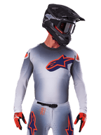 Alpinestars Supertech Lipan Jersey 2025, jersey de motocross de manga larga, gris con detalles en naranja y azul marino, camiseta técnica con perforaciones de ventilación, usado con casco negro y guantes rojos, diseño de competición estilizado