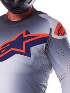 Primer plano de un piloto con el Jersey Alpinestars 2025 Supertech Lipan, jersey de motocross, gris y naranja flúor, con un gran logotipo naranja de Alpinestars en el pecho, perforaciones láser para ventilación y un casco negro, equipamiento de competición profesional.