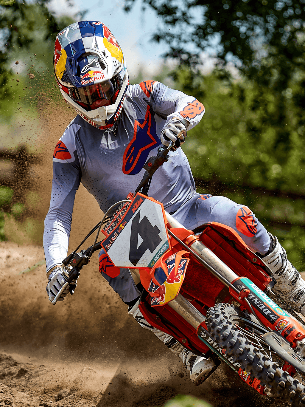 Piloto de motocross en acción en una pista de tierra vistiendo el jersey Alpinestars 2025 Supertech Lipan en gris naranja fluo, pantalones grises a juego con detalles naranjas, botas blancas y un casco de la marca Red Bull, tomando una curva en una moto de cross naranja con salpicaduras de tierra, equipamiento profesional de carreras todoterreno