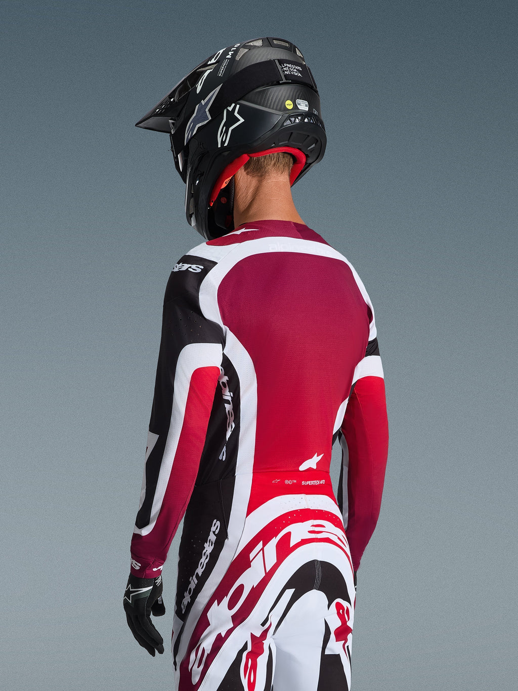 Vista trasera de un piloto de motocross con el Jersey Supertech AFD Vista 2026 en negro, rojo y blanco, combinado con pantalones a juego y un casco negro, con logotipos de Alpinestars destacados, paneles de malla rojos y un ajuste atlético de competición.