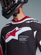 Alpinestars 2026 Supertech AFD Vista Jersey, primer plano de un piloto de motocross con un jersey negro con detalles en rojo y blanco, con un gran logotipo de Alpinestars en blanco y rojo, perforaciones de ventilación cortadas con láser y ajuste de competición profesional, combinado con un casco negro.