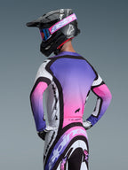 Piloto de motocross con el jersey Alpinestars 2026 Supertech AFD Vista en blanco, púrpura y negro, vista trasera que muestra un diseño con degradado de púrpura a rosa, combinado con pantalones a juego y casco negro, equipamiento profesional de carreras off-road.