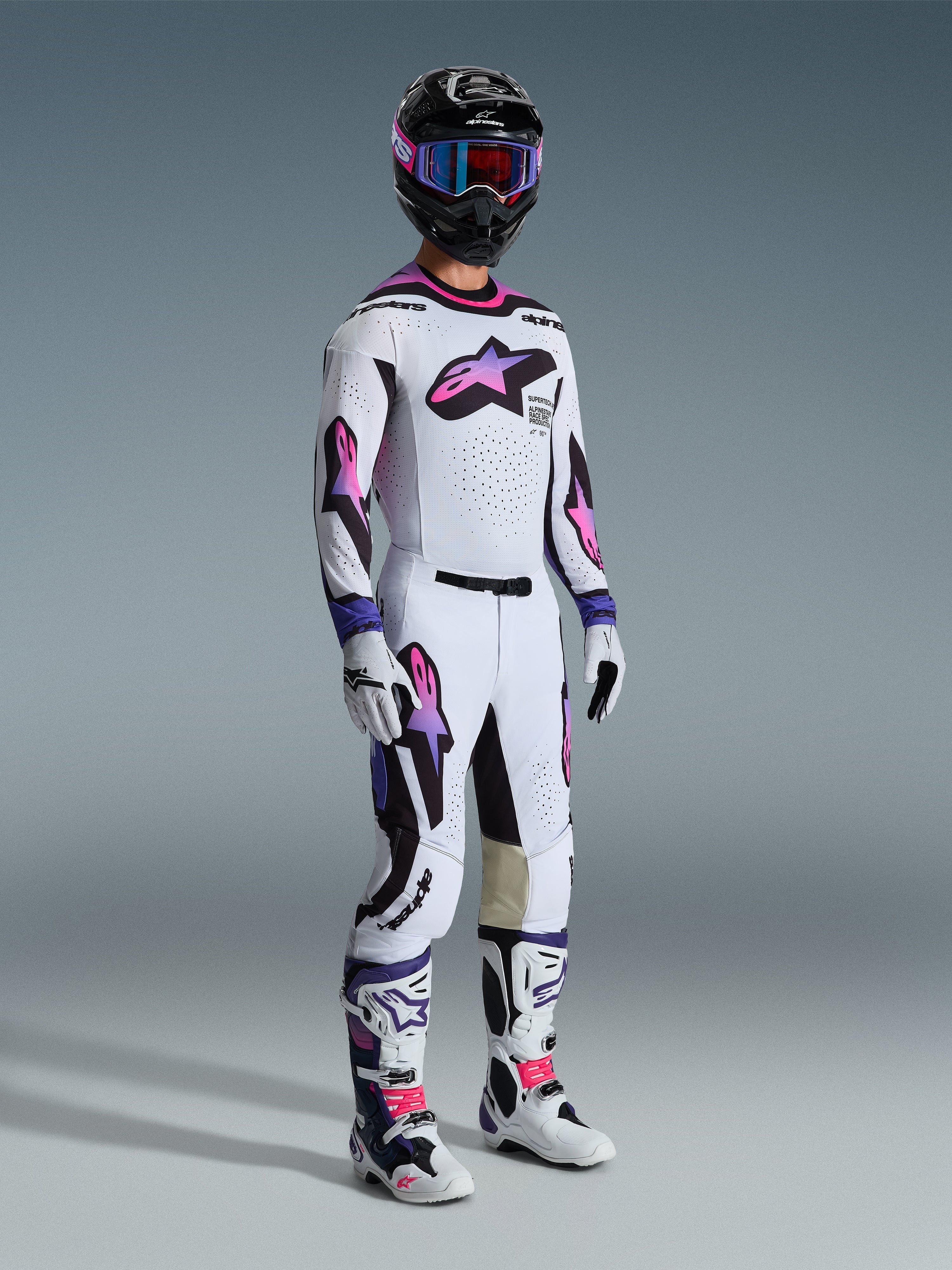 Piloto de motocross con el jersey Alpinestars 2026 Supertech AFD Vista en blanco, púrpura y negro, pantalones de motocross a juego con logotipos en degradado rosa y púrpura, casco negro con gafas púrpuras, botas blancas y negras y guantes blancos, conjunto completo de equipamiento profesional para carreras todoterreno