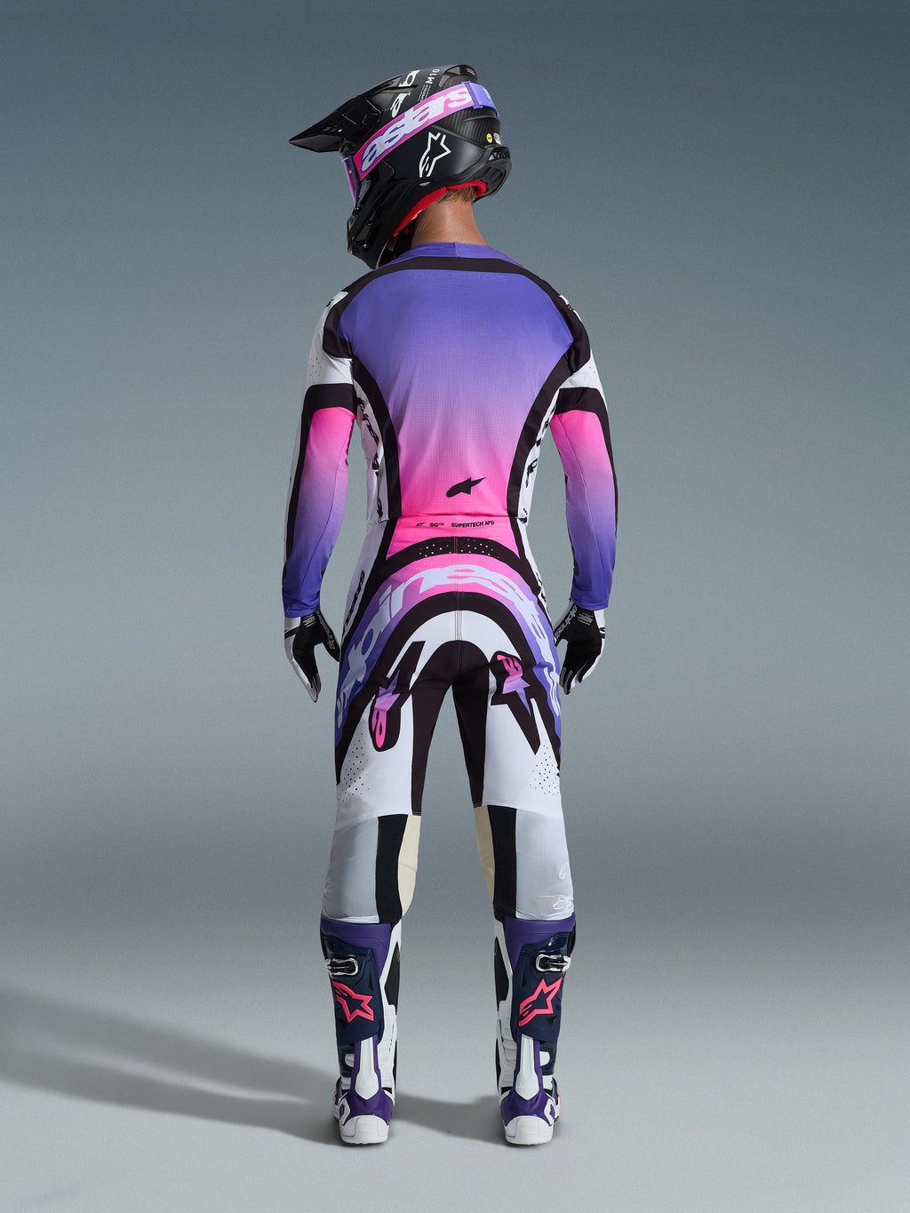 Piloto de motocross visto desde atrás con el Jersey Supertech AFD Vista 2026, blanco, morado y negro con detalles en degradado rosa, pantalones de motocross a juego, casco negro con gafas rosas y botas blancas y moradas, conjunto completo de equipamiento profesional de carreras