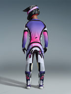 Piloto de motocross visto desde atrás con el Jersey Supertech AFD Vista 2026, blanco, morado y negro con detalles en degradado rosa, pantalones de motocross a juego, casco negro con gafas rosas y botas blancas y moradas, conjunto completo de equipamiento profesional de carreras