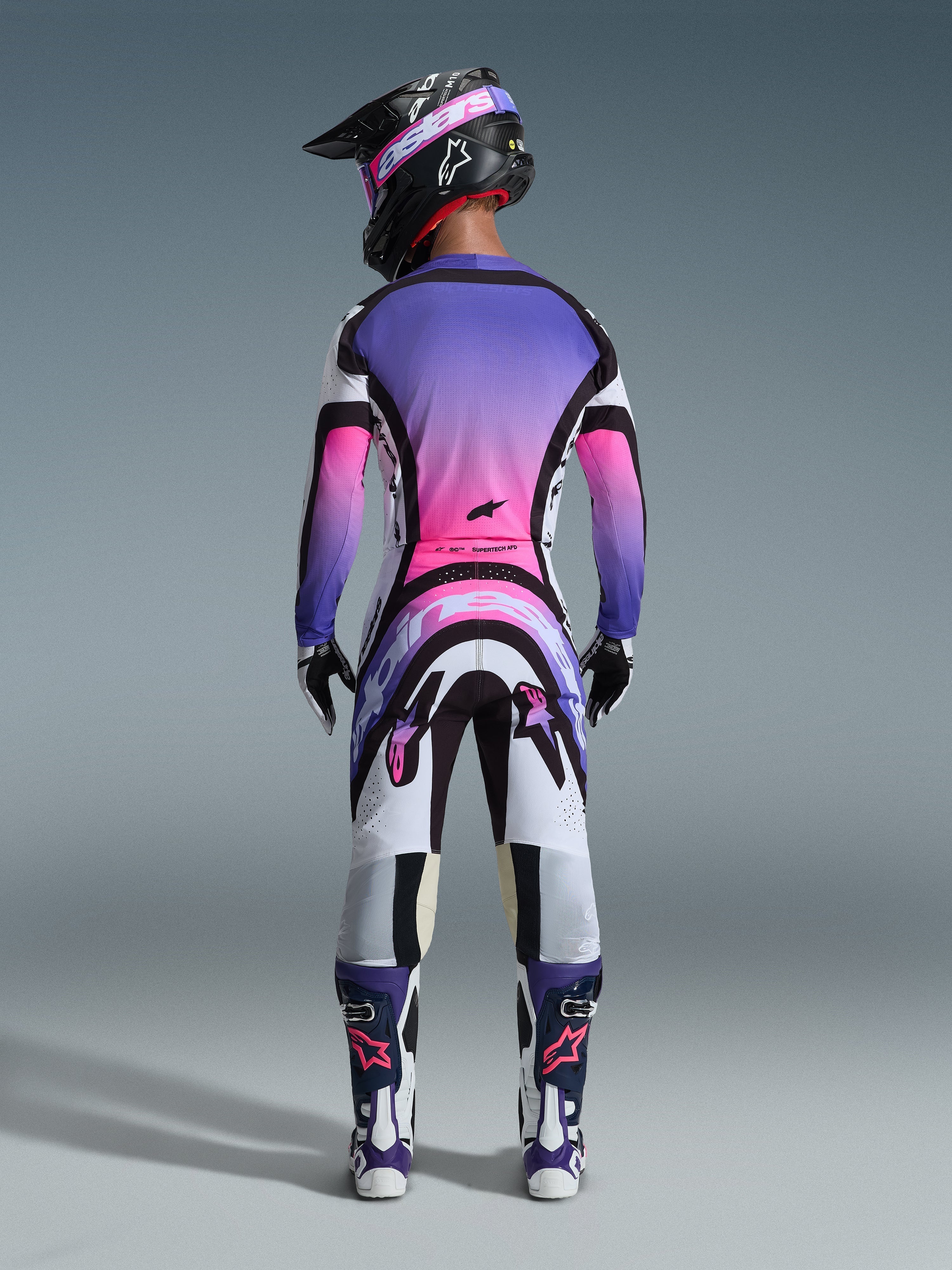 Piloto de motocross visto desde atrás con el Jersey Supertech AFD Vista 2026, blanco, morado y negro con detalles en degradado rosa, pantalones de motocross a juego, casco negro con gafas rosas y botas blancas y moradas, conjunto completo de equipamiento profesional de carreras