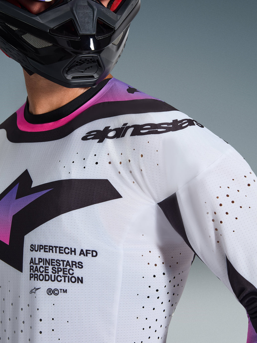 Alpinestars 2026 Supertech AFD Vista Jersey, jersey de motocross, blanco, morado y negro, vista de primer plano que muestra las perforaciones cortadas con láser y los logotipos de competición, usado con un casco negro, equipamiento profesional de carreras todoterreno