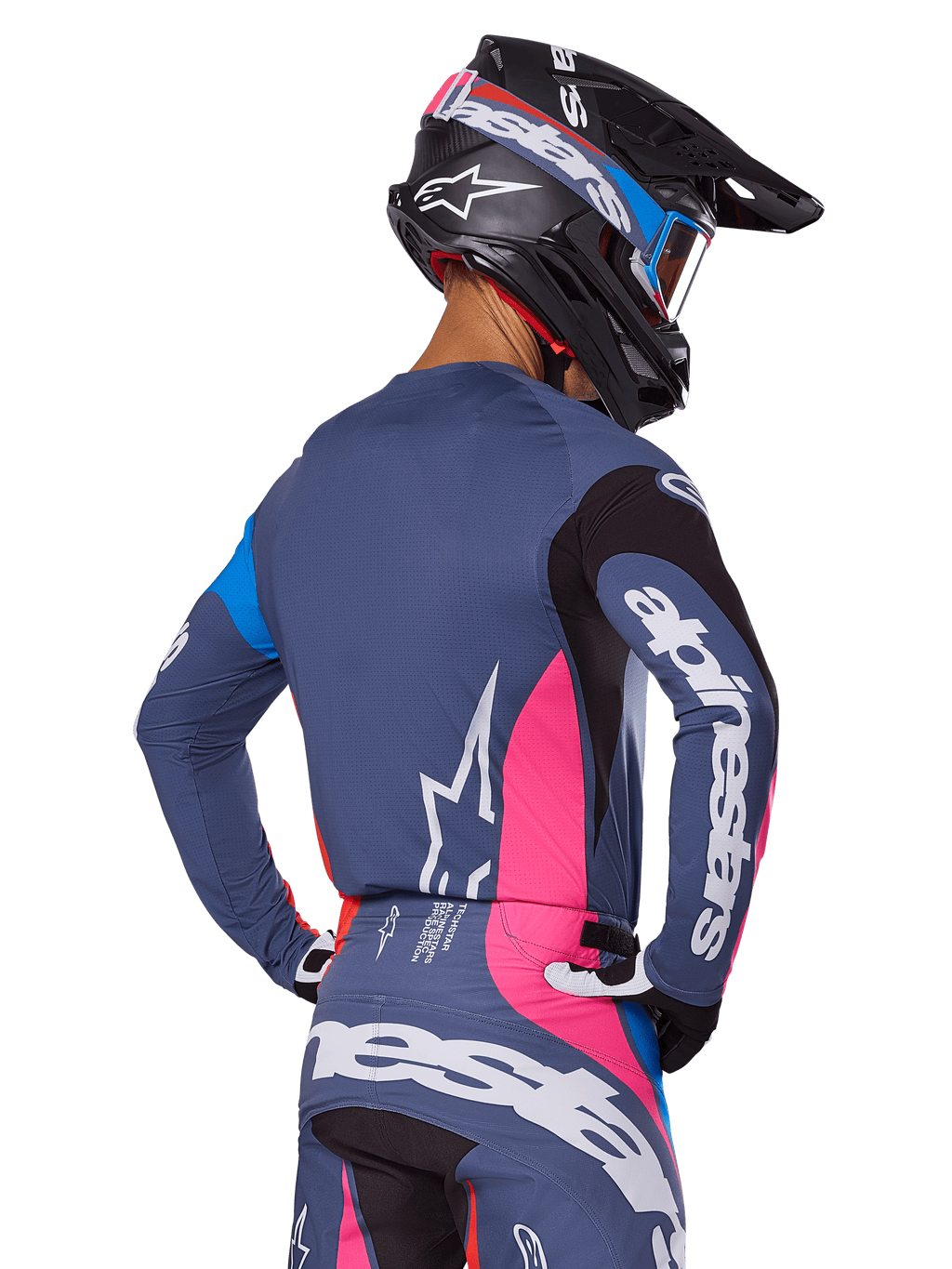 Piloto de motocross visto desde atrás vistiendo el conjunto de equipamiento Alpinestars 2025 Techstar Dreem en color gris, con un jersey y pantalones en gris azulado con paneles rosas y negros, logotipos blancos de Alpinestars en las mangas y la espalda, y un casco negro a juego con gráficos en azul y rosa.