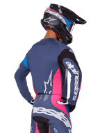 Piloto de motocross visto desde atrás vistiendo el conjunto de equipamiento Alpinestars 2025 Techstar Dreem en color gris, con un jersey y pantalones en gris azulado con paneles rosas y negros, logotipos blancos de Alpinestars en las mangas y la espalda, y un casco negro a juego con gráficos en azul y rosa.