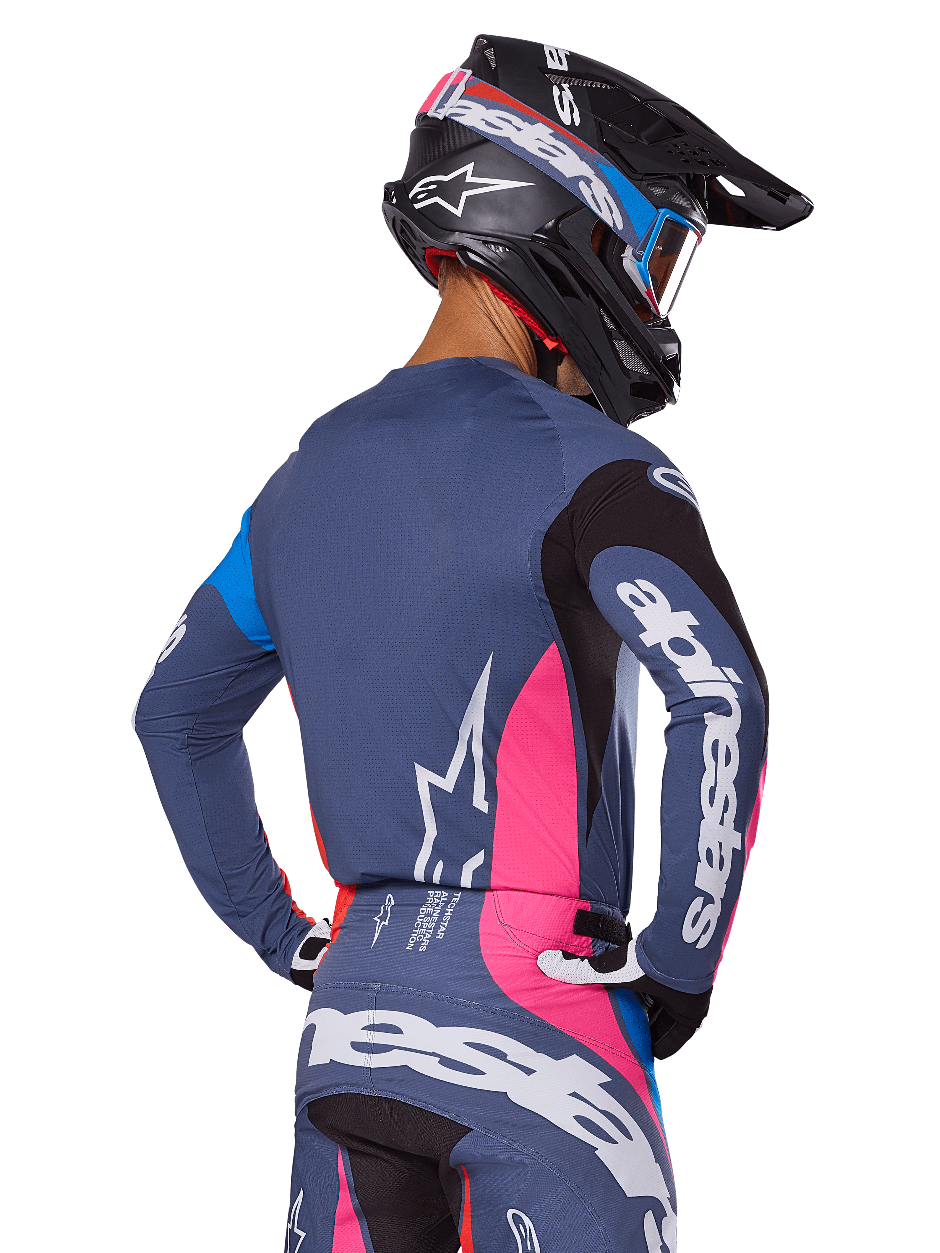 Piloto de motocross visto desde atrás vistiendo el conjunto de equipamiento Alpinestars 2025 Techstar Dreem en color gris, con un jersey y pantalones en gris azulado con paneles rosas y negros, logotipos blancos de Alpinestars en las mangas y la espalda, y un casco negro a juego con gráficos en azul y rosa.