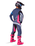 Piloto de motocross en vista trasera con el conjunto de equipamiento Alpinestars Techstar Dreem 2025, en gris con detalles en rosa vibrante y blanco, casco negro y blanco y botas rosa brillante, atuendo completo de carreras para motocross.