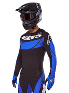 Piloto de motocross con el conjunto de equipo Alpinestars 2025 Techstar Dreem, jersey negro con paneles azules de estilo perforado y logotipos blancos, pantalones negros y azules a juego, casco de carreras negro y guantes azules, conjunto de alto rendimiento coordinado para motocross.