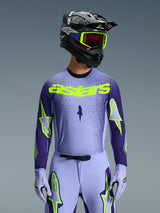 Alpinestars 2026 Supertech Scenz Jersey, jersey de motocross, morado y amarillo flúor, usado por un piloto con un casco negro y morado a juego, gafas amarillo neón y pantalones de carreras morados, con la marca astars en amarillo neón en el pecho y logos de estrellas en las mangas.