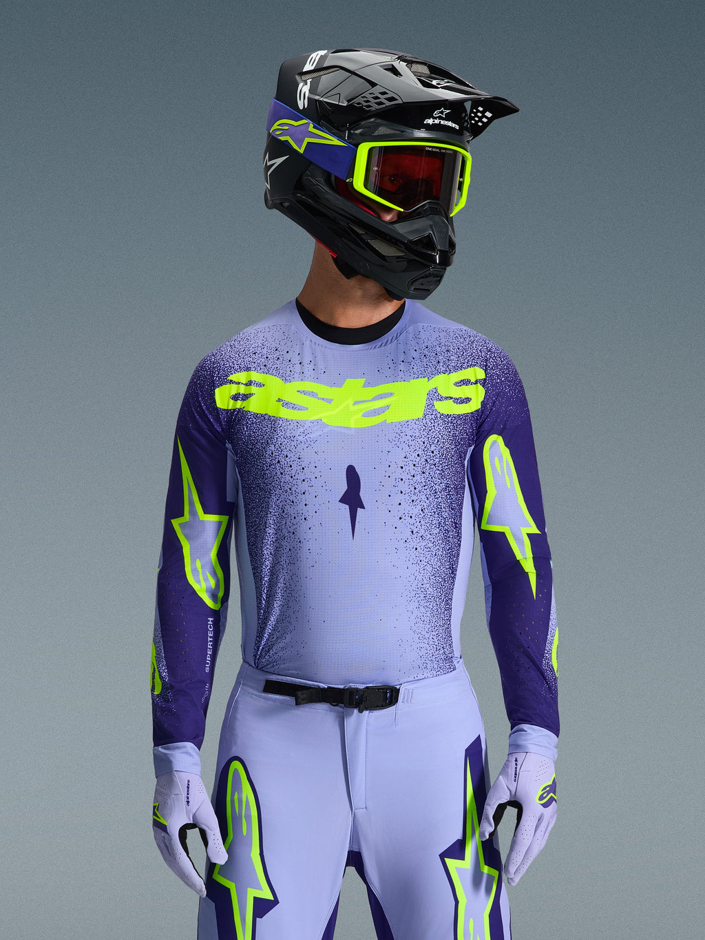 Frontal view of a motocross rider in the 2026 Supertech Scenz Gear Set, light purple and dark blue jersey with vibrant neon yellow 'astars' branding, matching light purple pants and gloves with neon accents, black helmet with neon yellow goggles, studio shot against a grey background
