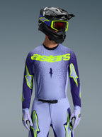 Frontal view of a motocross rider in the 2026 Supertech Scenz Gear Set, light purple and dark blue jersey with vibrant neon yellow 'astars' branding, matching light purple pants and gloves with neon accents, black helmet with neon yellow goggles, studio shot against a grey background