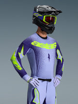 Piloto de motocross vistiendo el Jersey Alpinestars Supertech Scenz 2026, púrpura y amarillo flúor, pantalones púrpura a juego con detalles flúor, casco negro con gafas amarillas y guantes de carreras púrpura, equipamiento profesional coordinado para carreras off-road