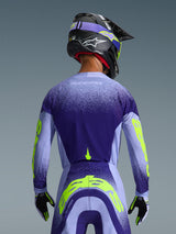 Alpinestars Supertech Scenz Jersey 2026 en púrpura y azul claro con detalles en amarillo fluorescente, vista trasera del piloto con pantalones de motocross a juego, casco negro y guantes, equipamiento deportivo de alto rendimiento para carreras todoterreno