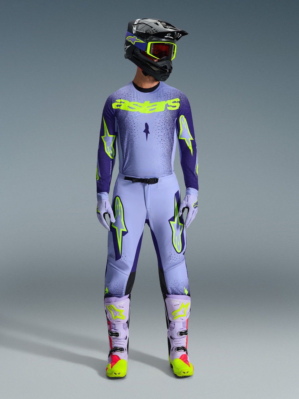 Motocross rider wearing complete Alpinestars 2026 Supertech Scenz racing gear, Purple and Yellow Fluo jersey with matching pants, black helmet with yellow goggles, light purple and neon boots, professional athletic-fit outfit for off-road racing