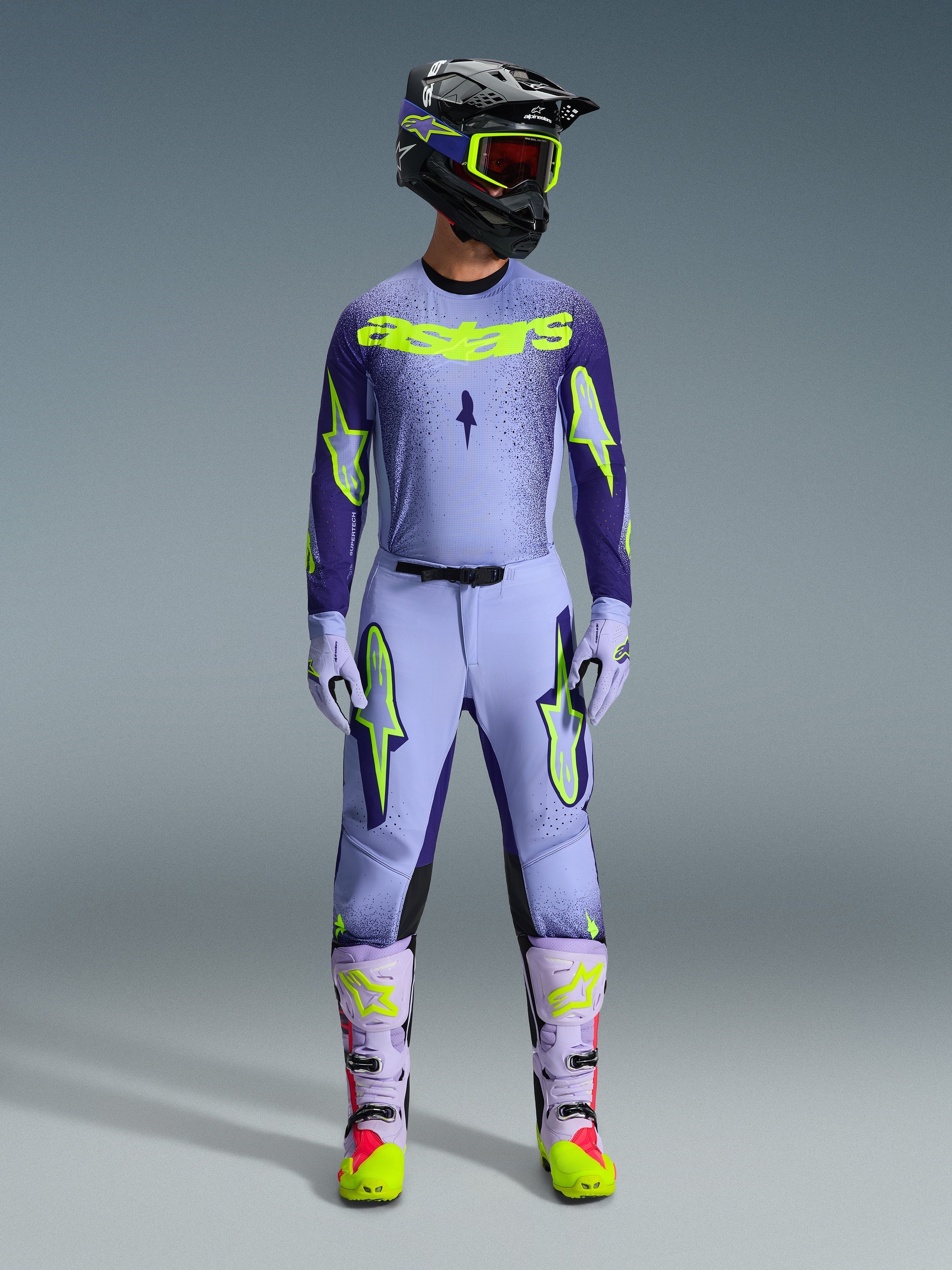 Motocross rider wearing complete Alpinestars 2026 Supertech Scenz racing gear, Purple and Yellow Fluo jersey with matching pants, black helmet with yellow goggles, light purple and neon boots, professional athletic-fit outfit for off-road racing