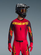 Alpinestars 2026 Supertech Scenz Jersey, jersey de motocross, rojo, negro y amarillo, usado por un piloto con casco negro a juego y guantes rojos, con un llamativo logotipo 'astars' amarillo en el pecho y un patrón de degradado moteado negro en los hombros.