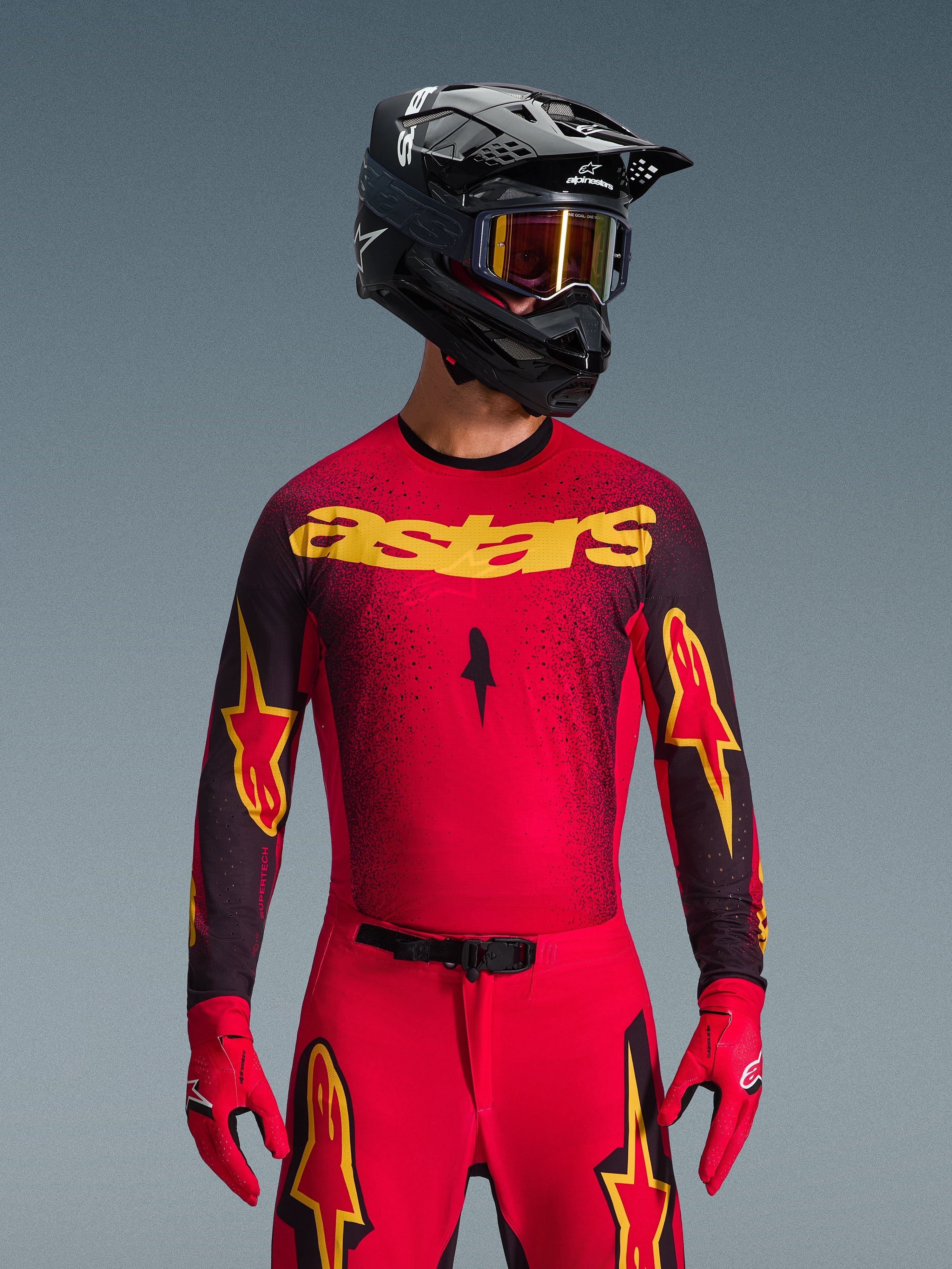 Alpinestars 2026 Supertech Scenz Jersey, jersey de motocross, rojo, negro y amarillo, usado por un piloto con casco negro a juego y guantes rojos, con un llamativo logotipo 'astars' amarillo en el pecho y un patrón de degradado moteado negro en los hombros.