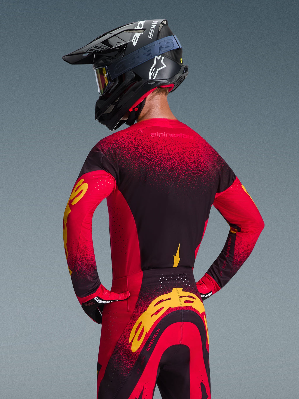 Alpinestars 2026 Supertech Scenz Jersey, rojo, negro y amarillo, vista trasera de un piloto de motocross con equipamiento de carreras a juego que incluye pantalones y casco negro, con un diseño degradado en rojo y negro y el llamativo logotipo de Alpinestars en amarillo