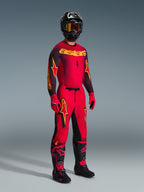 Piloto de motocross vistiendo el jersey Alpinestars Supertech Scenz 2026 en rojo, negro y amarillo, pantalones rojos a juego con detalles en negro y amarillo, casco negro con gafas, guantes rojos y botas negras con detalles rojos, conjunto completo de equipamiento de competición profesional.