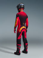 Piloto de motocross visto desde atrás con equipación completa de competición, jersey rojo con detalles en negro y amarillo, pantalones rojos y negros a juego con un gran logotipo amarillo de Alpinestars, casco negro, botas negras y rojas, guantes rojos y negros, conjunto coordinado para carreras profesionales de off-road