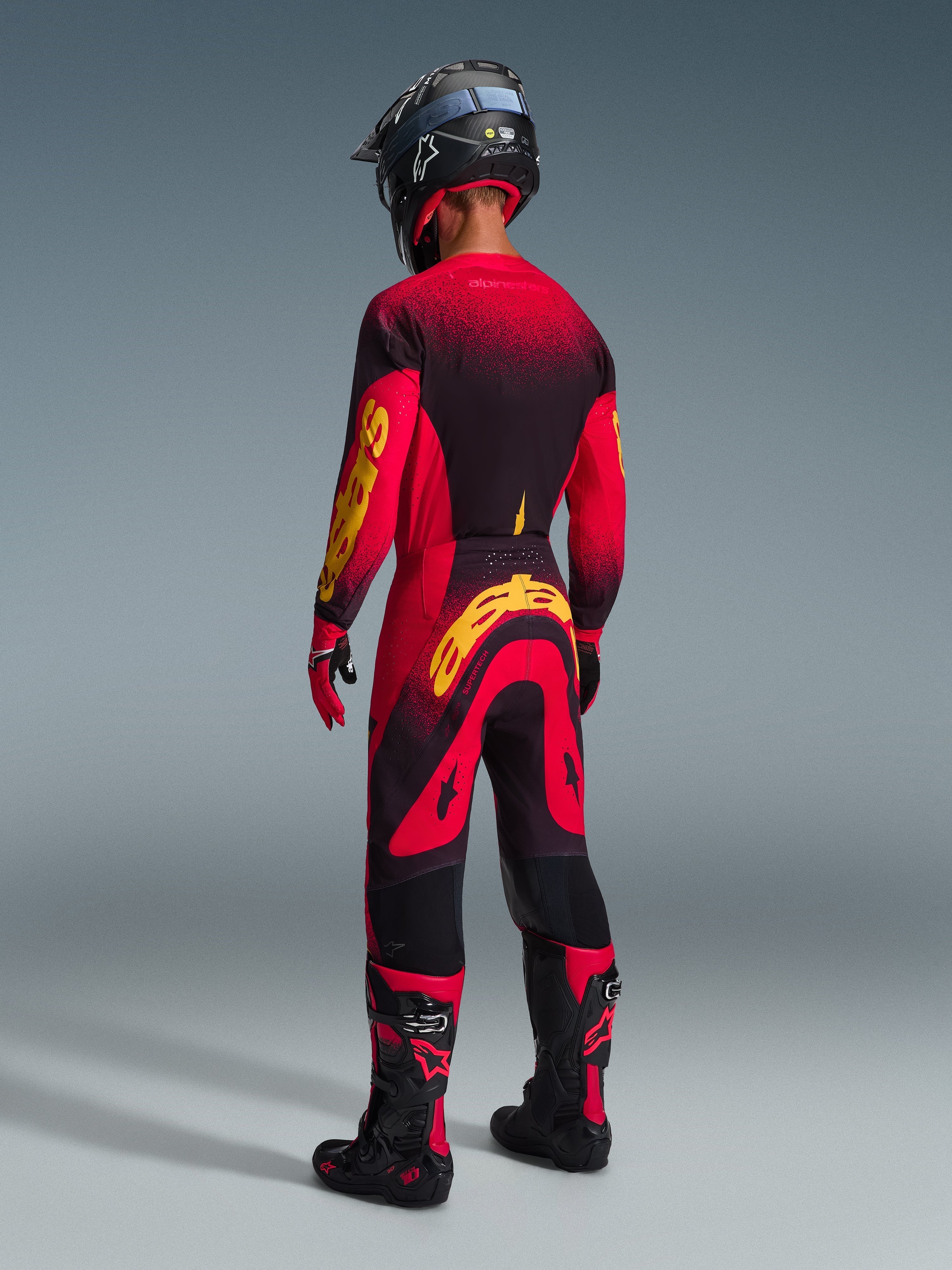 Piloto de motocross visto desde atrás con equipación completa de competición, jersey rojo con detalles en negro y amarillo, pantalones rojos y negros a juego con un gran logotipo amarillo de Alpinestars, casco negro, botas negras y rojas, guantes rojos y negros, conjunto coordinado para carreras profesionales de off-road