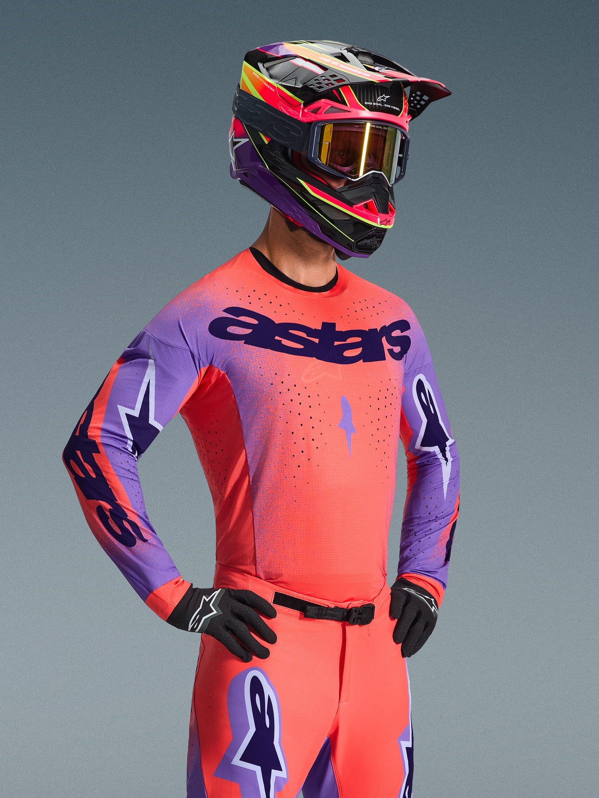 Motocross rider wearing the Alpinestars 2026 Supertech Scenz Jersey, Hot Coral and Purple with bold 'astars' chest branding, matching gear including coral pants and a coordinated purple and coral helmet, featuring laser-cut perforations for high-performance off-road racing.