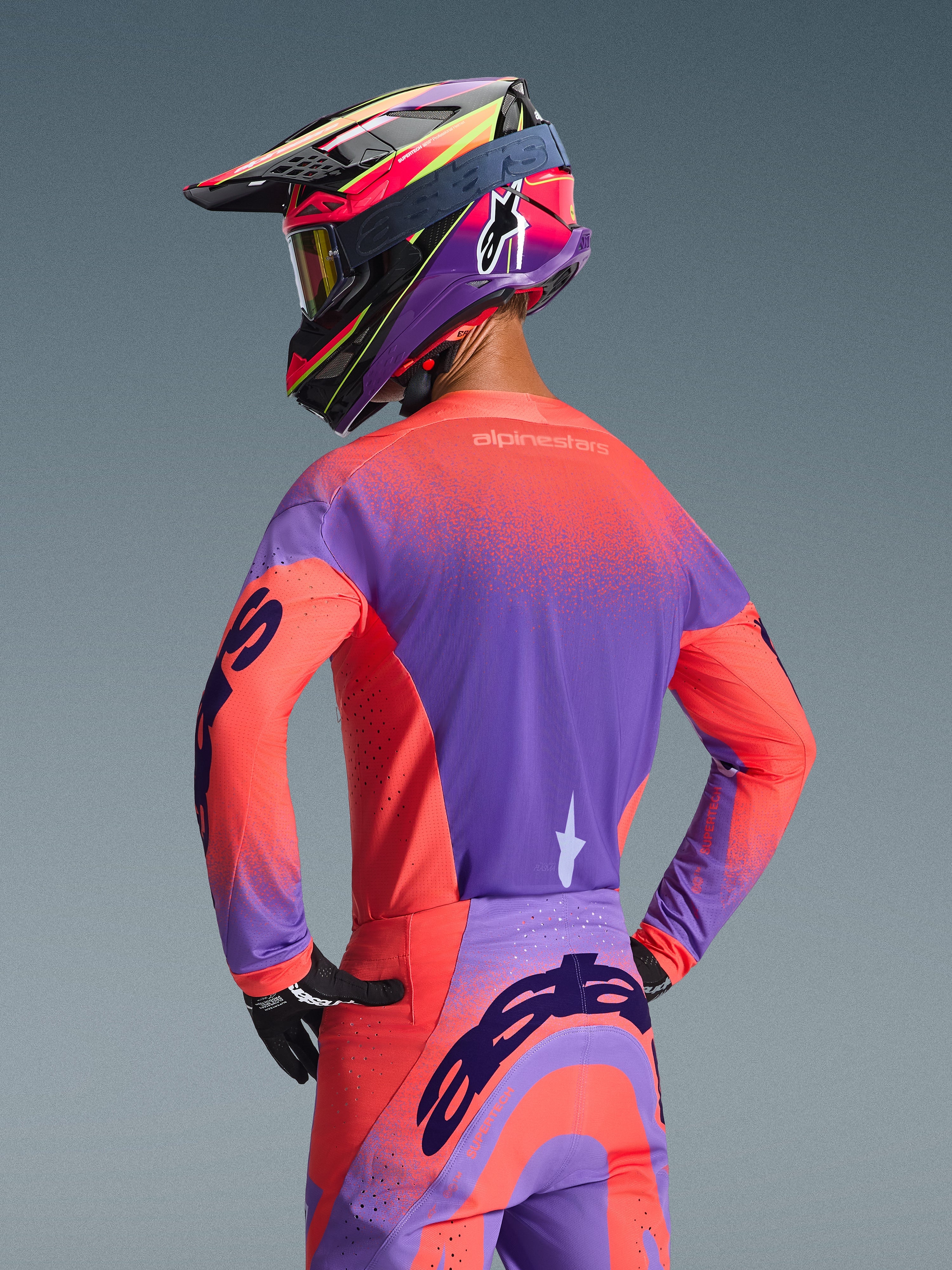Piloto de motocross visto desde atrás con el Jersey Alpinestars Supertech Scenz 2026 en color Hot Coral y púrpura, con un diseño de degradado moteado vibrante, combinado con pantalones de motocross a juego y un casco de competición colorido, equipamiento profesional para rendimiento off-road.
