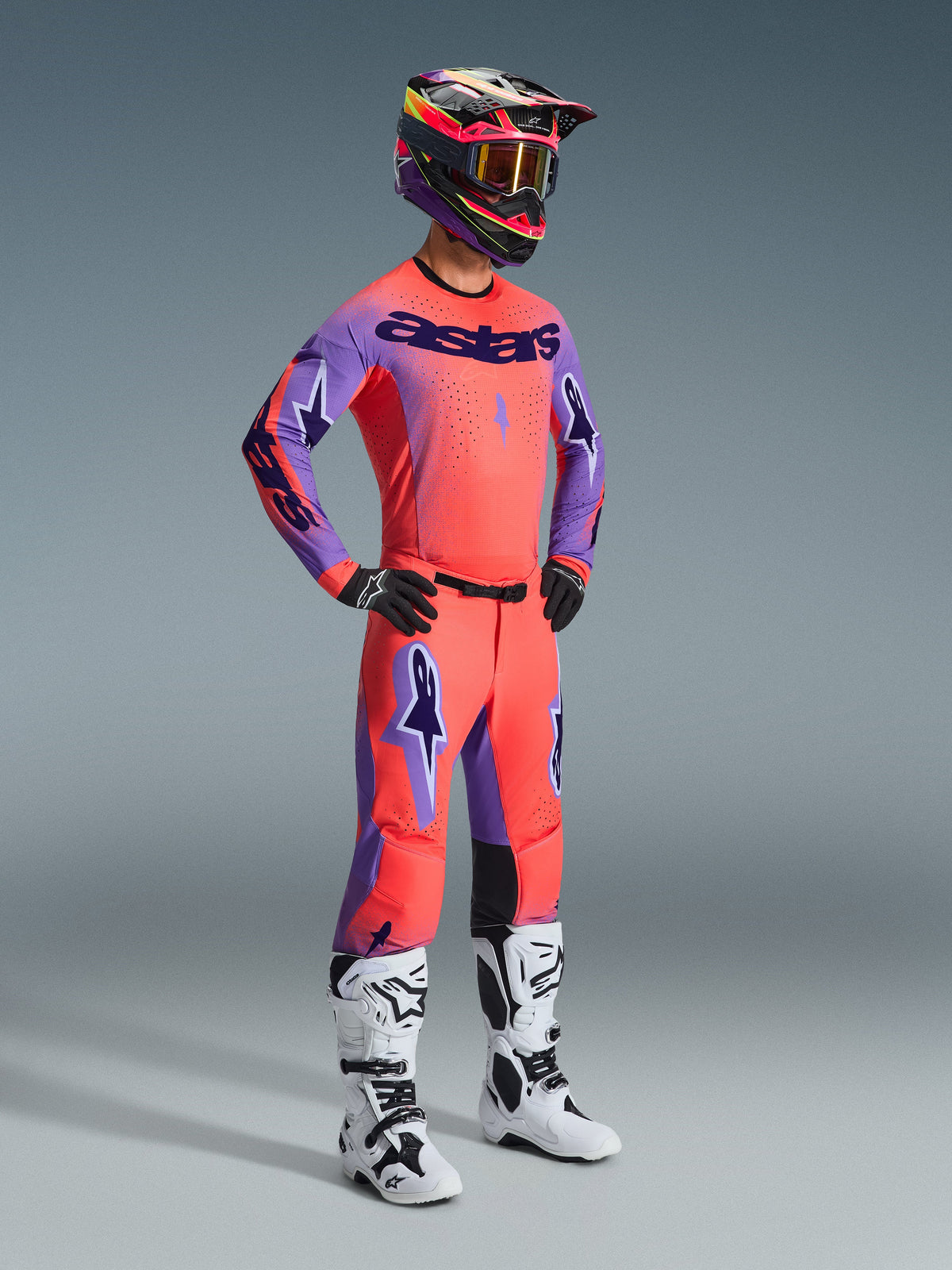 Motocross rider wearing complete Alpinestars 2026 Supertech Scenz Gear Set, Hot Coral jersey with purple sleeves and 'astars' branding, matching coral pants with purple accents, multi-colored helmet with gold-tinted goggles, white and black boots, black gloves, coordinated pro-level racing outfit for off-road performance