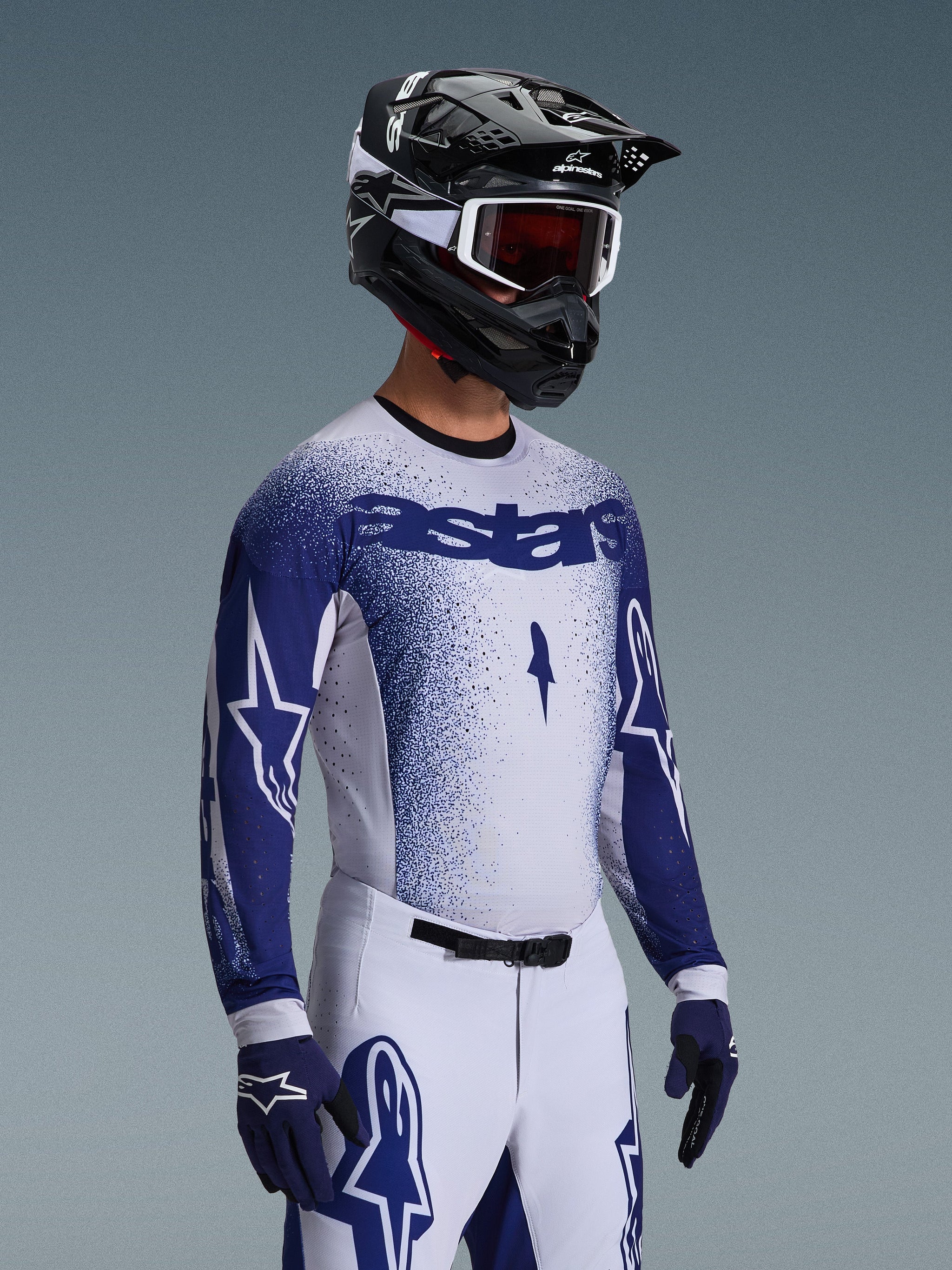 Piloto de motocross luciendo el Jersey Supertech Scenz 2026 en gris claro y azul oscuro, combinado con pantalones de carreras blancos y azules a juego, guantes azules y un casco negro con gafas blancas. El jersey presenta un efecto de degradado azul punteado en el pecho y las mangas, ajuste atlético profesional y el logotipo 'astars'.