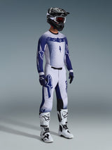 Piloto de motocross con equipamiento profesional completo, Jersey Alpinestars Supertech Scenz 2026 en gris claro y azul oscuro, pantalones de motocross a juego con gráficos de estrellas, casco negro con gafas rojas, botas de alto rendimiento en blanco y negro, y guantes coordinados en azul y blanco para competición.
