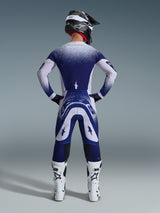 Piloto de motocross desde atrás con el Jersey Alpinestars Supertech Scenz 2026, gris claro y azul oscuro con un diseño de degradado punteado, pantalones de motocross a juego, botas de competición blancas y casco negro, equipamiento completo para carreras todoterreno.