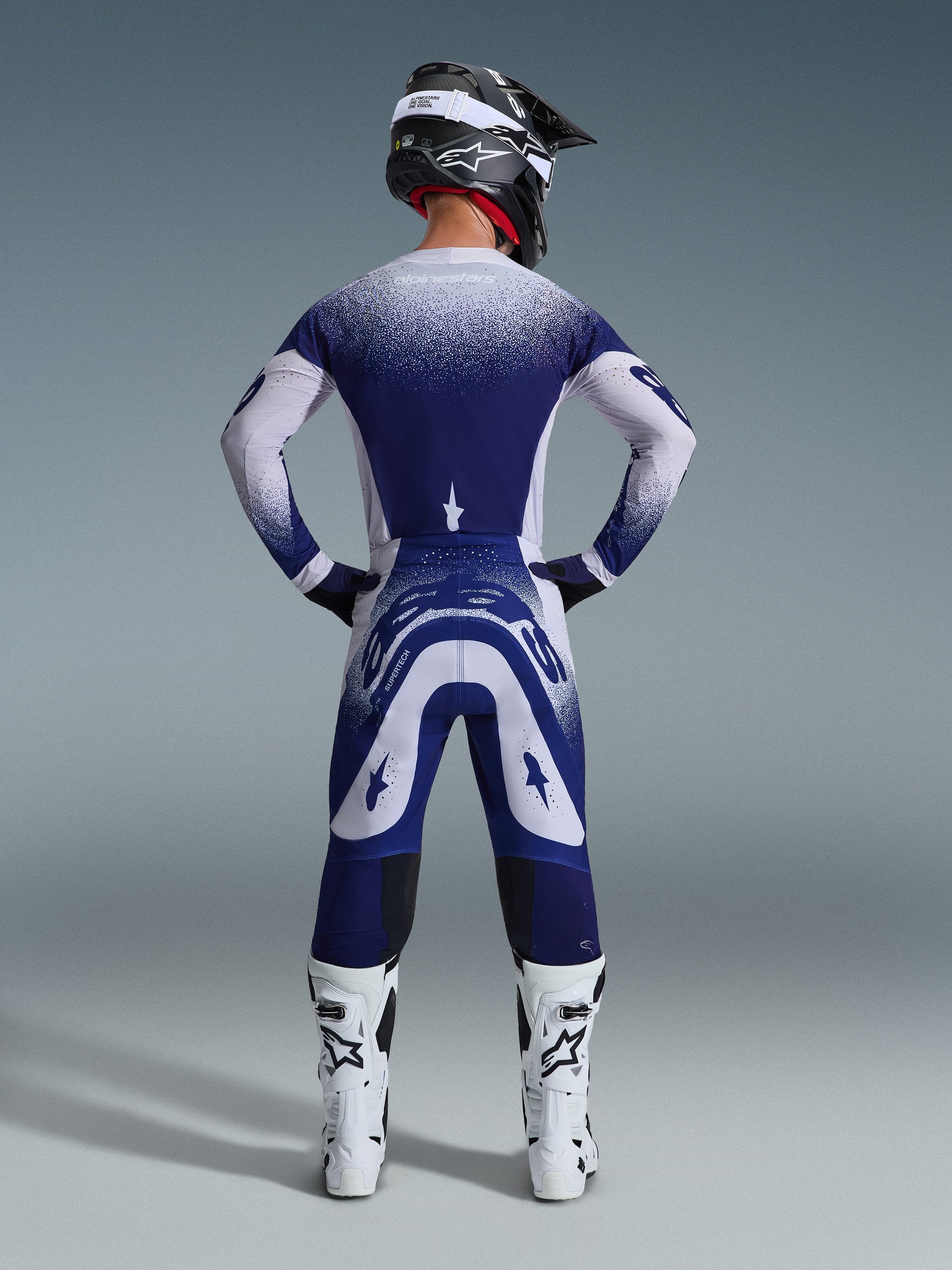 Piloto de motocross desde atrás con el Jersey Alpinestars Supertech Scenz 2026, gris claro y azul oscuro con un diseño de degradado punteado, pantalones de motocross a juego, botas de competición blancas y casco negro, equipamiento completo para carreras todoterreno.