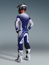 Vista trasera de un piloto de motocross con el Jersey Alpinestars Supertech Scenz 2026 en gris claro y azul oscuro, con un diseño de degradado moteado, combinado con pantalones a juego y botas de motocross blancas, equipamiento profesional de competición.