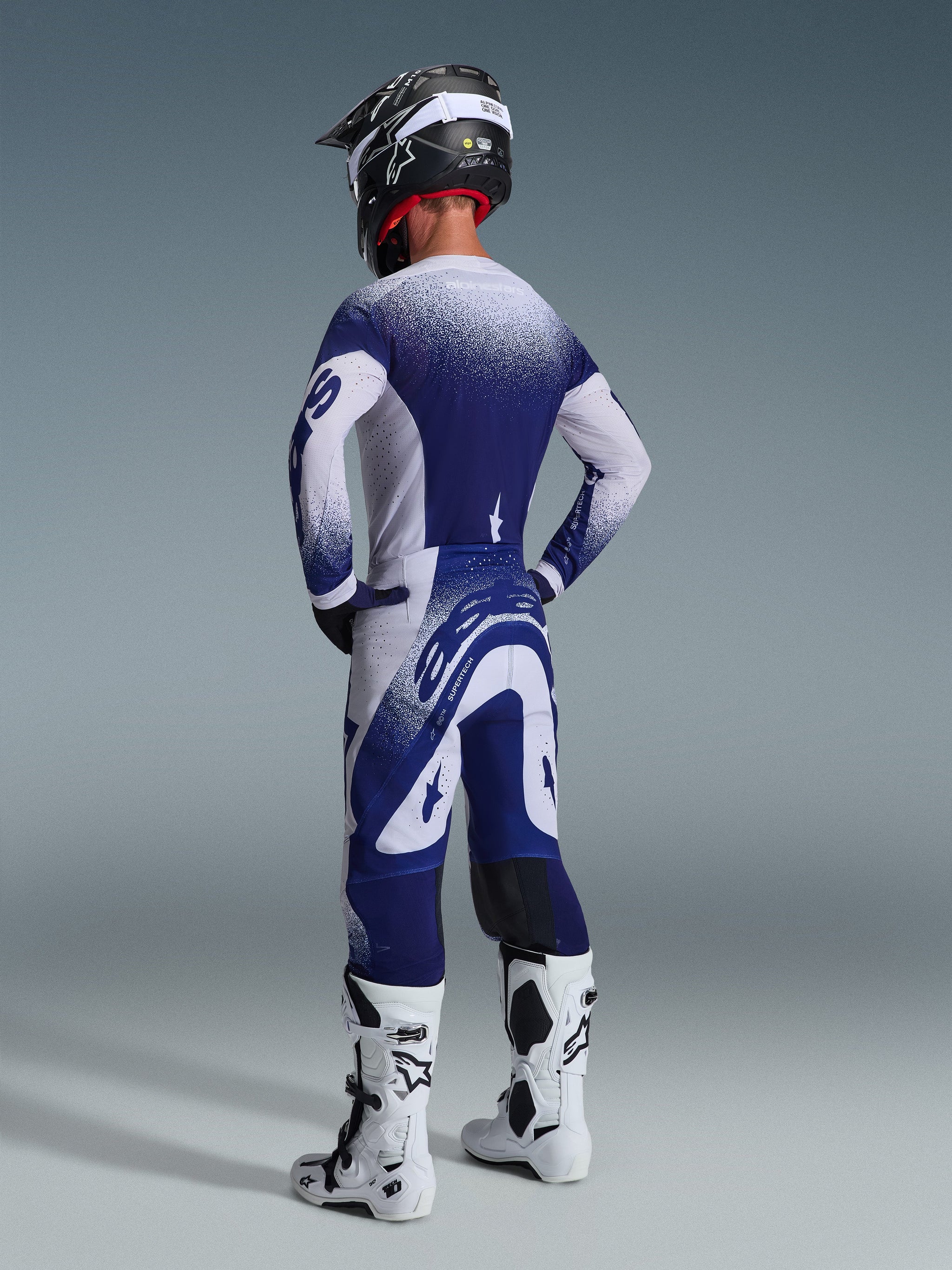 Vista trasera de un piloto de motocross con el Jersey Alpinestars Supertech Scenz 2026 en gris claro y azul oscuro, con un diseño de degradado moteado, combinado con pantalones a juego y botas de motocross blancas, equipamiento profesional de competición.