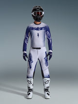 Piloto de motocross con equipamiento completo de competición, Jersey Supertech Scenz 2026 en gris claro y azul oscuro, pantalones de color gris claro a juego con logotipos azules, casco negro y blanco con gafas rojas, botas blancas y negras, guantes azul oscuro, conjunto coordinado para carreras de motocross