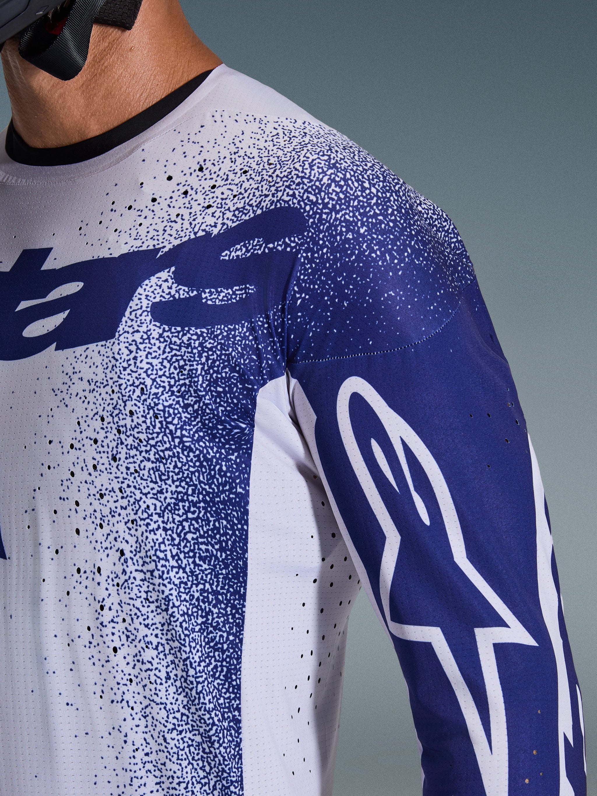 Primer plano del Jersey Alpinestars Supertech Scenz 2026 en gris claro y azul oscuro, con perforaciones cortadas con láser, patrón gráfico moteado, logotipo blanco de Astars en la manga, ajuste de rendimiento profesional para motocross
