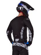 Piloto de motocross con el conjunto de equipo Alpinestars Racer Graphite 2025, vista posterior, jersey y pantalones negros con logotipos verticales de Alpinestars en gris, casco negro con detalles en azul y blanco, y guantes negros y azules, vestimenta coordinada para carreras de motocross