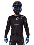 Alpinestars 2025 Racer Graphite Jersey, equipo de competición de motocross, negro con logotipos grises, usado con un casco negro a juego, gafas azules y guantes azules y grises, diseño de rendimiento ajustado con logotipos en el pecho y los brazos