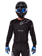 Alpinestars 2025 Racer Graphite Jersey, equipo de competición de motocross, negro con logotipos grises, usado con un casco negro a juego, gafas azules y guantes azules y grises, diseño de rendimiento ajustado con logotipos en el pecho y los brazos
