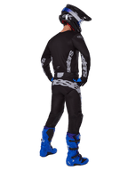 Piloto de motocross visto desde atrás con el conjunto Alpinestars 2025 Racer Graphite, jersey y pantalones negros y grises con logotipos grises, botas de motocross azul brillante, casco negro y azul, y guantes azules, equipamiento completo de alto rendimiento para off-road.