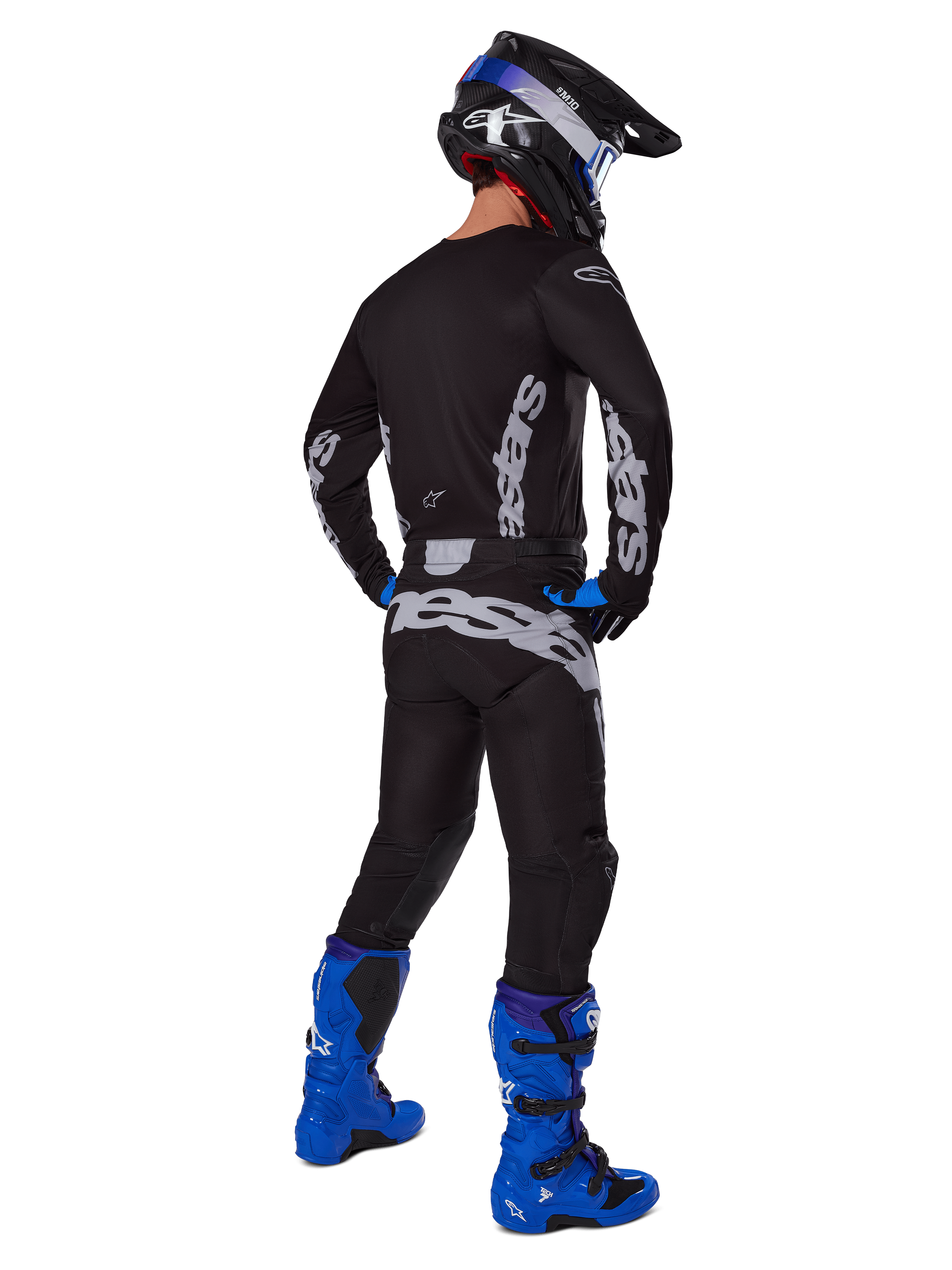 Piloto de motocross visto desde atrás con el conjunto Alpinestars 2025 Racer Graphite, jersey y pantalones negros y grises con logotipos grises, botas de motocross azul brillante, casco negro y azul, y guantes azules, equipamiento completo de alto rendimiento para off-road.