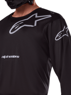 Alpinestars 2025 Racer Graphite Jersey, negro y gris, primer plano de piloto con jersey de motocross, con marca Alpinestars y logotipos A-star en gris, material de politejido técnico para conducción fuera de carretera