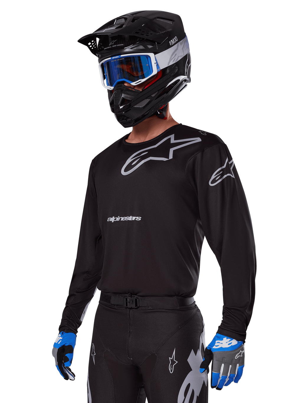 Piloto de motocross con el conjunto de equipo Alpinestars 2025 Racer Graphite, jersey negro con logotipos Alpinestars en gris, pantalones negros a juego con detalles grises, casco negro con gafas azules y blancas, guantes azules y grises, atuendo técnico estilizado para carreras fuera de carretera