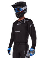 Piloto de motocross con el conjunto de equipo Alpinestars 2025 Racer Graphite, jersey negro con logotipos Alpinestars en gris, pantalones negros a juego con detalles grises, casco negro con gafas azules y blancas, guantes azules y grises, atuendo técnico estilizado para carreras fuera de carretera